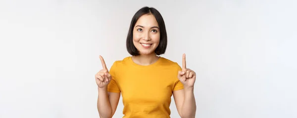 Šťastná krásná asijská dívka ukazuje reklamu, ukazuje prsty nahoru, stojící nad bílým pozadím — Stock fotografie