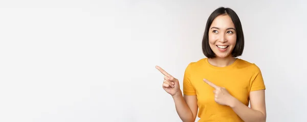 Retrato de chica morena asiática sonriente en camiseta amarilla, señalando los dedos a la izquierda, mostrando espacio para copiar, trato promocional, pancarta demostrativa, de pie sobre fondo blanco — Foto de Stock