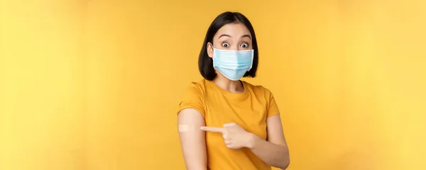 Covid ve sağlık konseptinden aşı. Tıbbi yüz maskesi takmış heyecanlı Asyalı kadın, sarı arka planda dikilen yara bandıyla parmağını omzuna doğrultuyor. — Stok fotoğraf