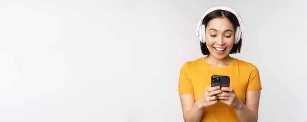 Jolie fille japonaise dans les écouteurs, regardant le téléphone mobile et souriant, en utilisant l'application de musique sur smartphone, debout sur fond blanc — Photo
