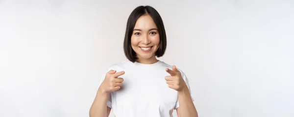 照片上的亚洲女孩微笑着用手指对着相机，挑选，邀请你，祝贺你，站在白色背景的T恤上 — 图库照片