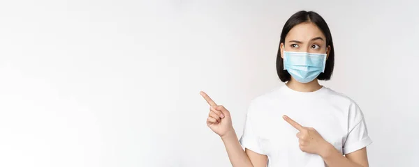 Изображение азиатки в медицинской маске из шабаша, выглядящей смущенно слева, указывающей на логотип, показывающей рекламу, стоящей на белом фоне — стоковое фото