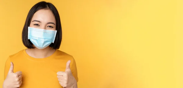 Mulher coreana alegre em máscara facial médica, apoiar as pessoas durante a pandemia, usar equipamentos de proteção individual de covid-19, mostrando polegares para cima na aprovação, fundo amarelo — Fotografia de Stock