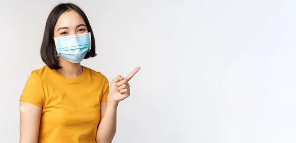 Covid-19, vaccinatie en gezondheidszorg concept. Portret van schattig aziatisch meisje met medisch masker, heeft pleister op de schouder na coronavirus vaccin, staande over witte achtergrond — Stockfoto