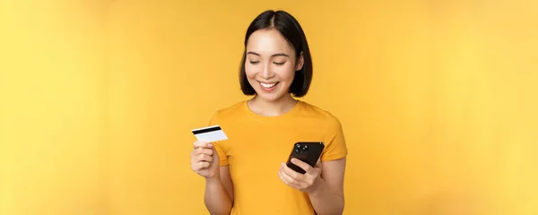 Compras en línea. Sonriendo chica asiática utilizando la tarjeta de crédito y la aplicación de teléfono móvil, pagar sin contacto, orden en la aplicación de teléfono inteligente, de pie sobre fondo amarillo — Foto de Stock