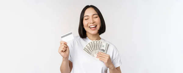 Portrait of asian woman smiling, holding credit card and money cash, dollars, standing in tshirt over white background — Stok Foto