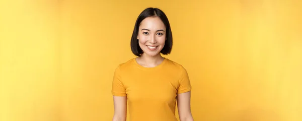 Retrato de jovem mulher asiática moderna, sorrindo feliz com dentes brancos, olhando confiante para a câmera, vestindo t-shirt casual, de pé sobre fundo amarelo — Fotografia de Stock