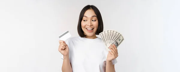 Portrait of asian woman smiling, holding credit card and money cash, dollars, standing in tshirt over white background — Stock Photo, Image