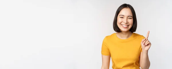 Hermosa joven asiática mujer apuntando con el dedo hacia arriba, sonriendo y mirando divertido a la cámara, mostrando publicidad, anuncio en la parte superior, fondo blanco —  Fotos de Stock