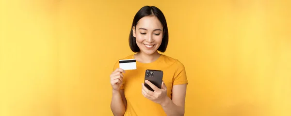 Compras en línea. Sonriendo chica asiática utilizando la tarjeta de crédito y la aplicación de teléfono móvil, pagar sin contacto, orden en la aplicación de teléfono inteligente, de pie sobre fondo amarillo — Foto de Stock