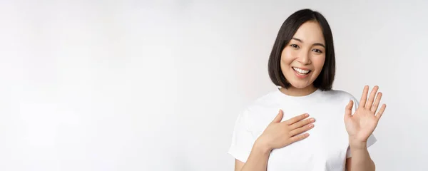 Retrato de hermosa chica coreana levantando la mano, presentarse, poner la mano en el corazón, saludo, de pie sobre fondo blanco —  Fotos de Stock