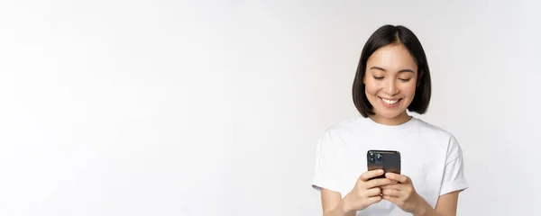 Porträtt av leende asiatisk kvinna med hjälp av mobiltelefon, chatta, sms: a meddelande, står i t-shirt över vit bakgrund — Stockfoto