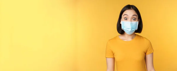 Portret van Koreaans meisje in medisch gezichtsmasker kijken verrast, verbaasd reactie op nieuws, staande over gele achtergrond — Stockfoto
