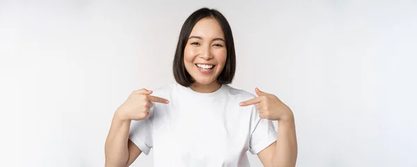 행복하고 자신감 있는 동양 여성, 웃으며 자신을 가리키고, 자신을 홍보하고, 티셔츠에 로고를 표시하고, 하얀 배경 위에 서서 — 스톡 사진
