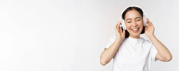 Nowoczesna azjatycka dziewczyna tańczy, słucha muzyki ze słuchawkami, uśmiecha się szczęśliwy, stojąc w koszulce na białym tle — Zdjęcie stockowe