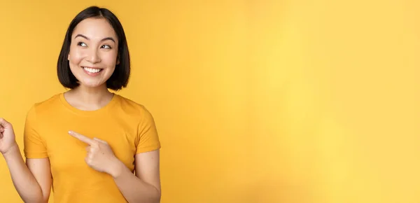Retrato de chica asiática feliz señalando los dedos y mirando a la izquierda, sonriendo asombrado, mirando banner promocional, mostrando publicidad sobre fondo amarillo — Foto de Stock