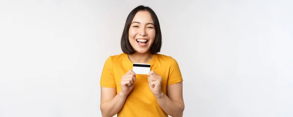 Retrato de hermosa chica coreana sosteniendo tarjeta de crédito, recomendando servicio bancario, de pie en camiseta amarilla sobre fondo blanco —  Fotos de Stock