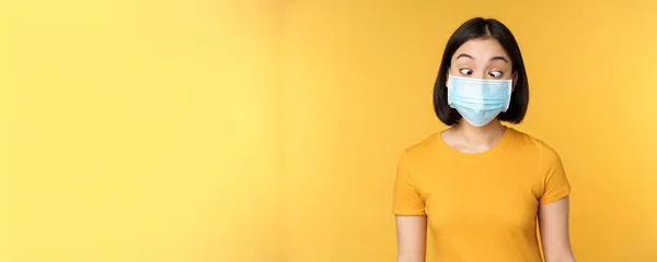 Concepto de salud y personas. Funny asiática chica entrecerrando los ojos, mirando su cara máscara médica, de pie sobre fondo amarillo —  Fotos de Stock