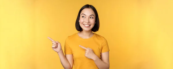 一个快乐的亚洲女孩的画像，手指指向左边，面带微笑，惊讶地看着横幅，在黄色的背景下展示广告 — 图库照片