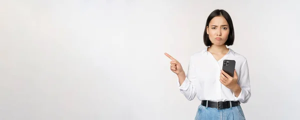 Skeptical asian modern girl, holding smartphone, pointing finger left at empty space and frowing hesitant, has doubts, standing over white background — Stock Photo, Image