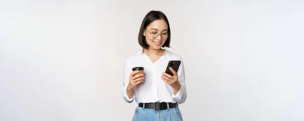 Bild av modern asiatisk kvinna tittar på mobiltelefon, dricka hämtmat kaffe, bär glasögon, med hjälp av smartphone app, står över vit bakgrund — Stockfoto