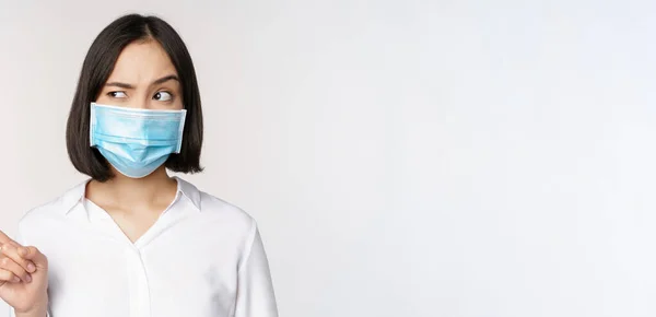 Bild av ung asiatisk kvinna i medicinsk ansiktsmask, pekar vänster och ser med misstänksam förvirrad uttryck, står över vit bakgrund — Stockfoto