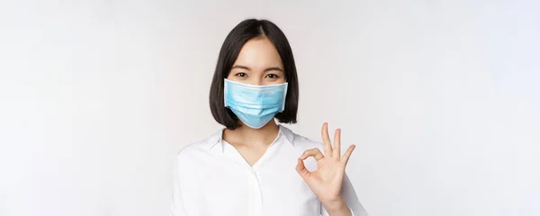 Covid och hälsa koncept. Porträtt av asiatisk kvinna bär medicinsk ansiktsmask och visar okej tecken, står över vit bakgrund — Stockfoto