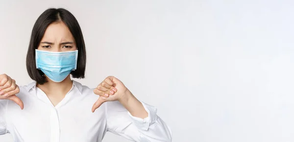 Porträtt av asiatisk kvinna i medicinsk ansiktsmask visar tummen ner med besviken, trött ansiktsuttryck, står över vit bakgrund — Stockfoto