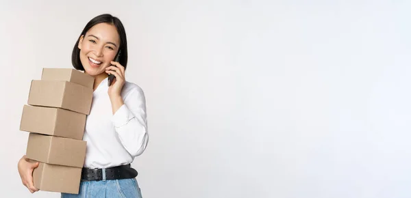 Imagen de joven mujer de negocios asiática contesta llamada telefónica mientras lleva cajas para la entrega, posando sobre fondo blanco —  Fotos de Stock