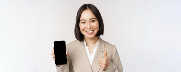 Mujer asiática sonriente mostrando la pantalla del teléfono inteligente y pulgares hacia arriba. Persona corporativa demuestra interfaz de aplicación de teléfono móvil, de pie sobre fondo blanco — Foto de Stock