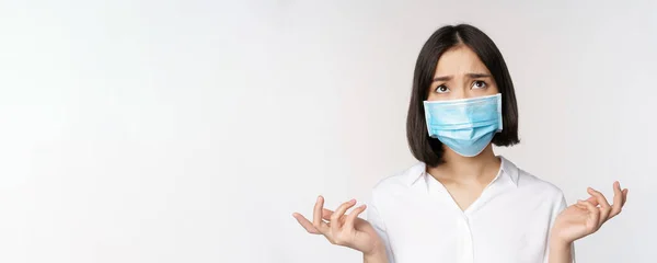 Angustiada y miserable joven asiática mujer en la máscara de la cara, mirando hacia arriba, mirando hacia arriba triste, de pie sobre fondo blanco —  Fotos de Stock