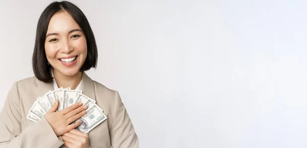 Glad asiatisk affärskvinna innehar kontanter, kramar dollar pengar och ler, står över vit bakgrund i kostym — Stockfoto