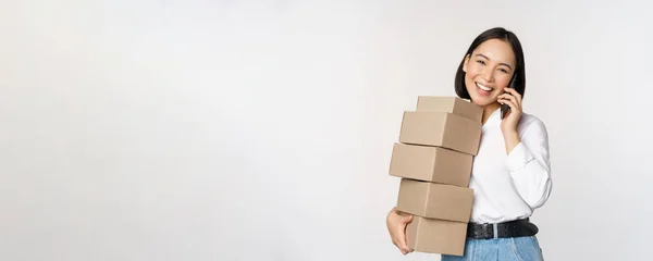 Imagem de jovem mulher de negócios asiática atender telefonema enquanto carrega caixas para entrega, posando contra fundo branco — Fotografia de Stock