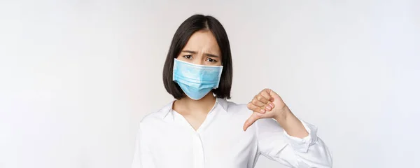 Porträtt av asiatisk kvinna i medicinsk ansiktsmask visar tummen ner med besviken, trött ansiktsuttryck, står över vit bakgrund — Stockfoto