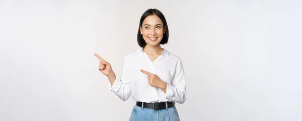 Portrait d'une femme asiatique attirante pointant vers la gauche avec un sourire heureux, montrant la bannière ou le logo de côté, debout sur fond de studio blanc — Photo