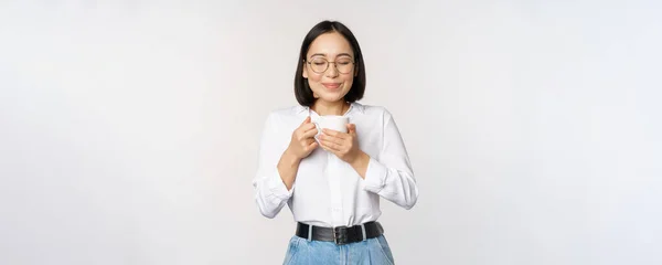 笑顔の韓国人女性が嗅ぎ、マグカップでコーヒーの匂いを嗅ぎ、おいしい飲み物を楽しみ、白い背景の上に立つイメージ — ストック写真
