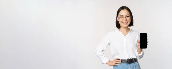 Smiling korean woman showing smartphone screen, demonstrating mobile application, standing over white background — Stock Photo, Image