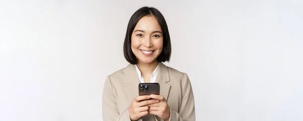 Bild av asiatisk affärskvinna i kostym, hålla mobiltelefon, med hjälp av smartphone app, ler mot kameran, vit bakgrund — Stockfoto