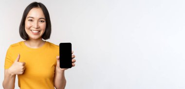 Teknoloji ve insan konsepti. Gülümseyen genç bir kadın başparmağını kaldırıyor ve akıllı telefon ekranı, cep telefonu uygulama arayüzü, beyaz arka plana karşı duruyor.