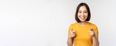 Genç Koreli kız öğrenci, kamerayı işaret ediyor ve gülümsüyor, seni tebrik ediyor, insanları seçiyor, beyaz arka planda duruyor.