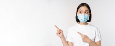 Tıbbi maske takan genç Koreli kadın solu işaret ediyor ve logoya bakıyor, reklam ya da afiş gösteriyor, beyaz arka planda duruyor.