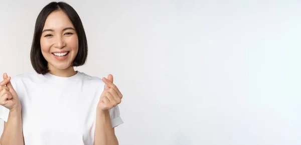 Schöne asiatische Frau lächelt, zeigt Fingerherzen Geste, trägt T-Shirt, steht vor weißem Hintergrund — Stockfoto