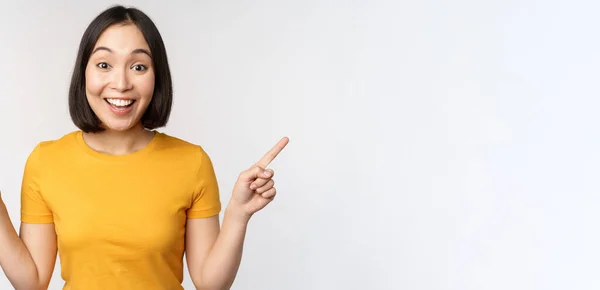 Linda chica asiática señalando los dedos hacia los lados, mostrando promo izquierda y derecha, dos opciones, variantes de productos, de pie en camiseta amarilla sobre fondo blanco — Foto de Stock