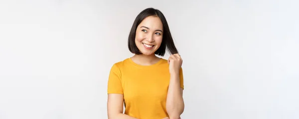 Красива романтична азіатська дівчина, посміхається і грає з волоссям, дивлячись щасливо на камеру, стоїть в жовтій футболці на білому тлі — стокове фото