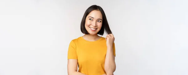 Linda menina asiática romântica, sorrindo e brincando com o cabelo, olhando feliz para a câmera, de pé em camiseta amarela sobre fundo branco — Fotografia de Stock