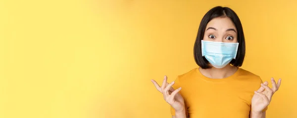 Covid-19 en medisch concept. Close-up portret van aziatische vrouw in het gezicht masker, op zoek verrast en verbaasd over nieuws, staande over gele achtergrond — Stockfoto