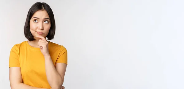 Imagem de pensar mulher asiática, olhando para o lado e ponderando, tomada de decisão, de pé em camiseta amarela sobre fundo branco — Fotografia de Stock