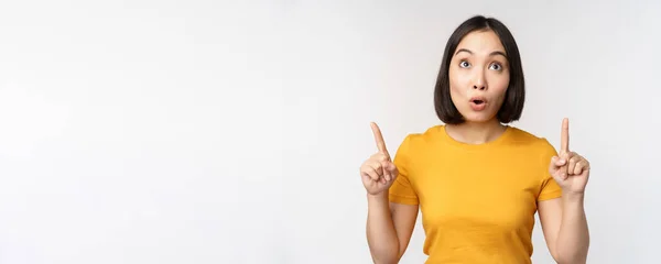 Gelukkig mooi aziatisch meisje tonen advertentie, wijzend vingers omhoog, staande over witte achtergrond — Stockfoto
