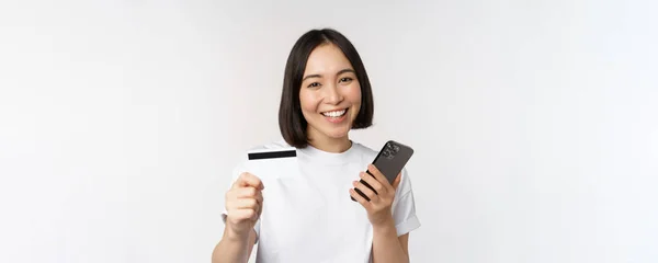 Усміхнена азіатська дівчина використовує мобільний телефон, показує кредитну картку, концепцію безконтактних платежів, онлайн-покупки, стоїть на білому тлі — стокове фото