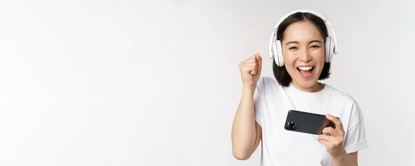 Felice ragazza asiatica gamer, giocando sul cellulare, guardando su smartphone, indossando le cuffie, in piedi su sfondo bianco — Foto Stock